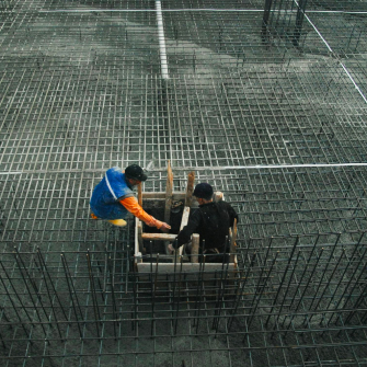 A construction site with rebar layed out on the ground and two workers. One of the workers is descending down a ladder and into a hole.